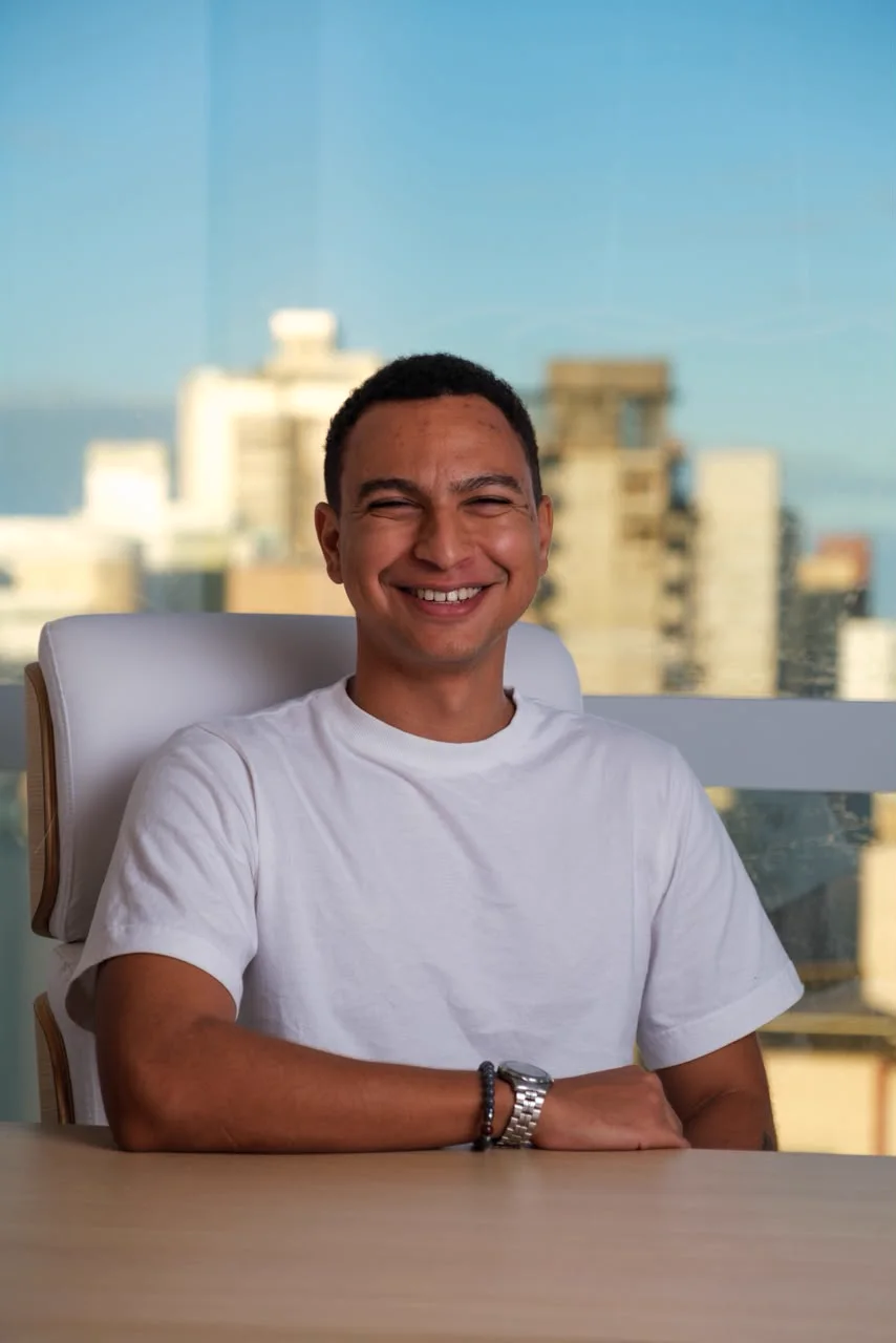 Yury Sixel sorrindo, sentado à mesa do consultório, com a cidade ao fundo